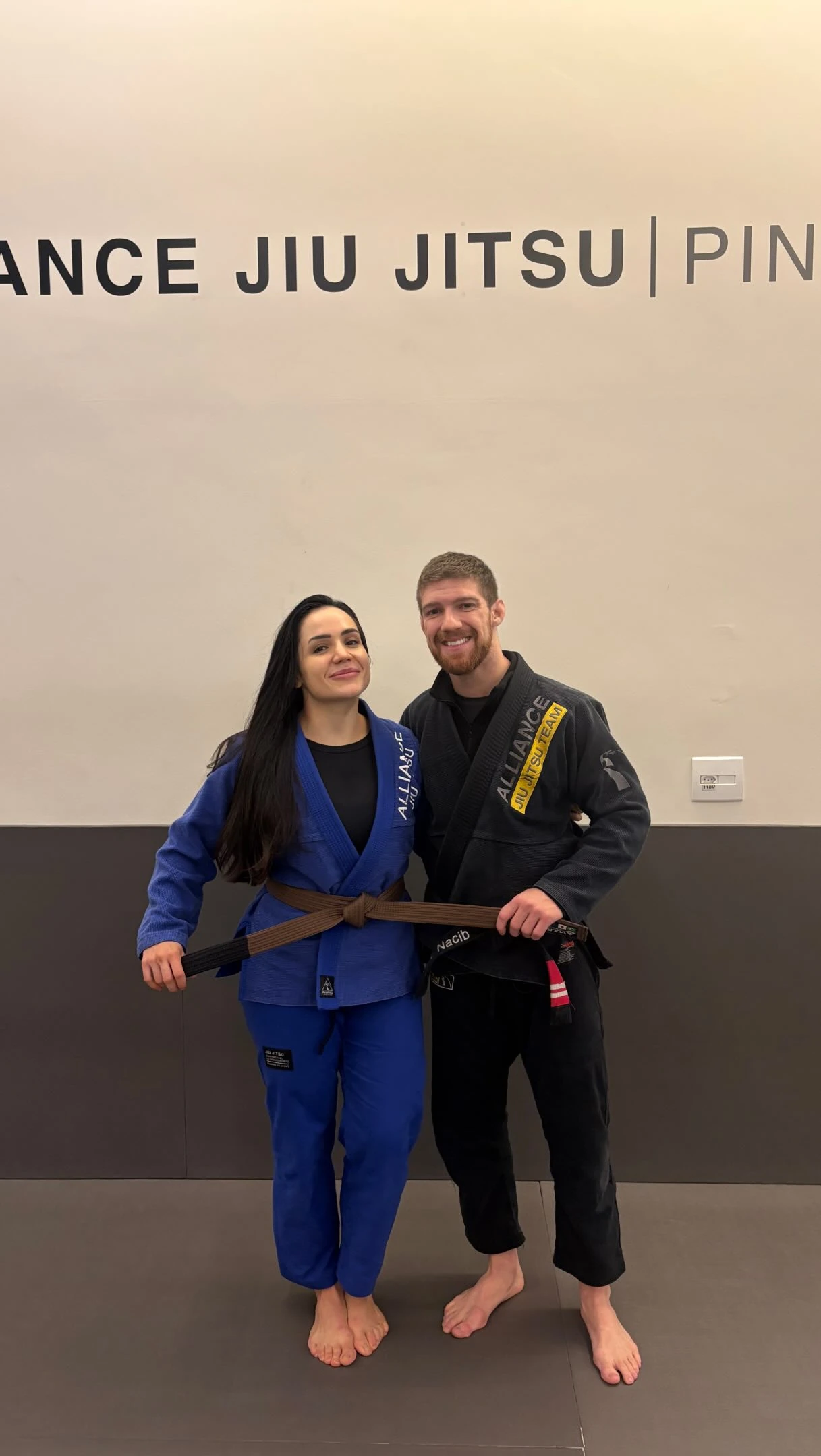 Fernanda Cosi sendo promovida à faixa marrom de jiu-jitsu ao lado de um amigo de equipe